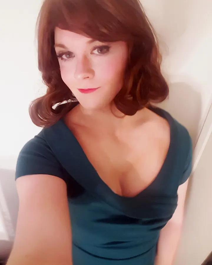 Femme trans européenne aux cheveux roux courts, en robe verte ajustée avec décolleté profond, regardant directement l'objectif avec un sourire discret.