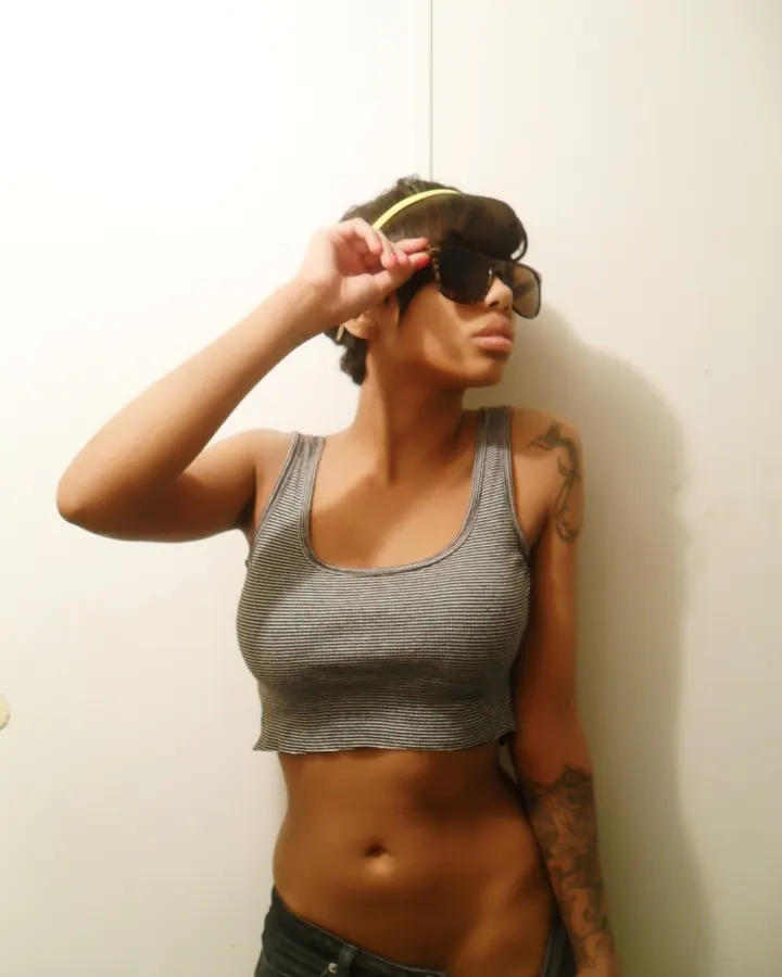 Femme trans latino aux cheveux courts noirs, en crop-top gris rayé et jean, portant des lunettes de soleil et tenant une main sur son front, avec tatouages sur le bras.