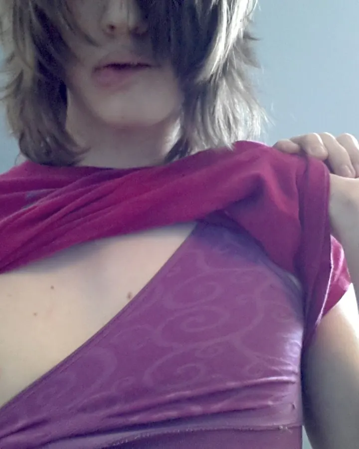 Femme trans aux cheveux bruns, portant un haut violet, regardant l'objectif dans un cadre intérieur.