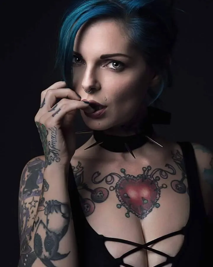Jeune femme trans européenne aux cheveux gris, portant une robe violette, assise sur un lit entourée de peluches, main dans les cheveux.