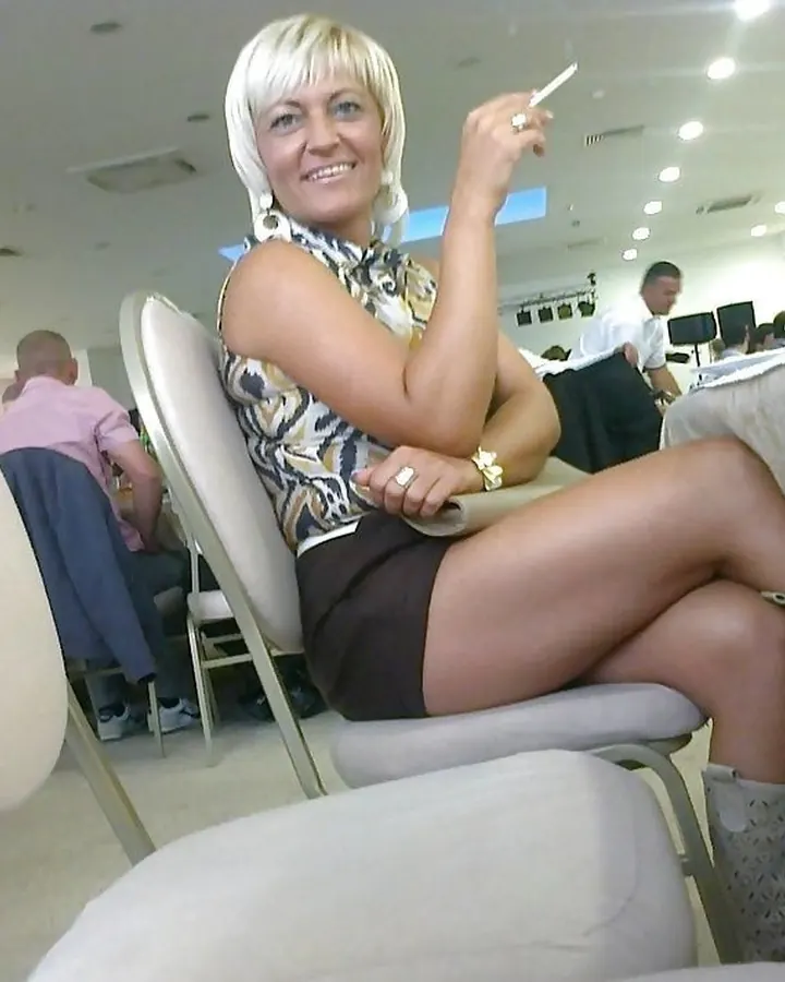 Femme trans aux cheveux blonds courts en robe violette, assise sur une chaise dans un restaurant, tenant une cigarette et souriant.