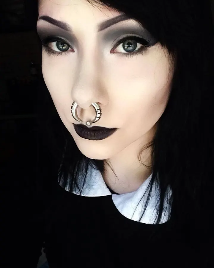 Femme trans aux cheveux noirs longs en lingerie noire, posant de face avec des chaînes autour du cou, montrant ses tatouages.