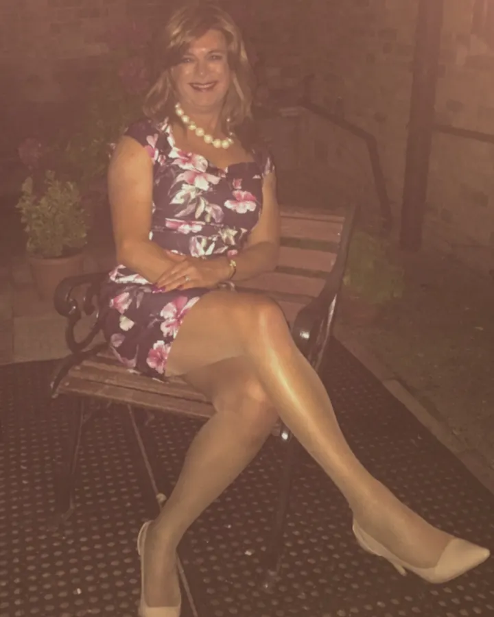 Femme trans aux cheveux blonds, souriante, en robe fleurie, assise sur un banc en extérieur, portant des chaussures à talons clairs.