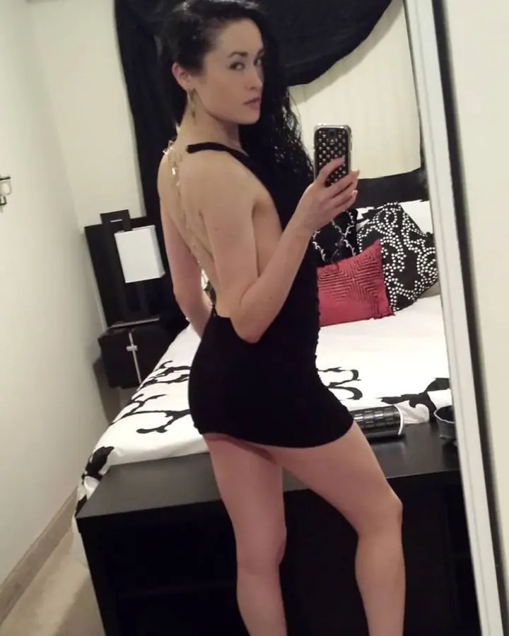 Femme trans aux cheveux noirs portant une robe noire à dos nu, prenant un selfie devant un miroir dans une chambre.